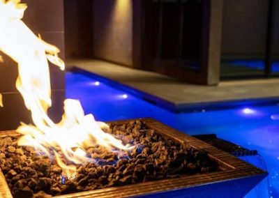 Fire pit with bright flames beside a lit swimming pool at night, creating a cozy and inviting outdoor atmosphere.