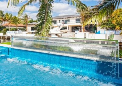 Infinity acrylic swimming pool with a glass waterfall and palm trees overlooking a luxurious villa under a clear blue sky.