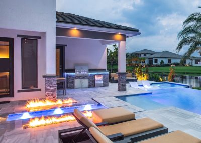 Luxurious outdoor kitchen with modern fire pit, pool, and lounge chairs at dusk.
