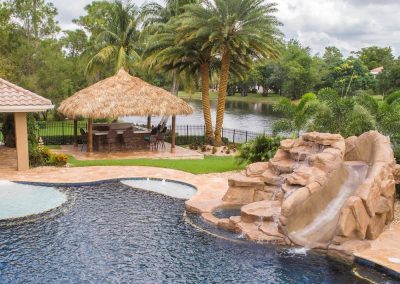 Luxury backyard pool with waterfall, tiki hut, and lush greenery by a lake. Perfect tropical escape and relaxation spot.