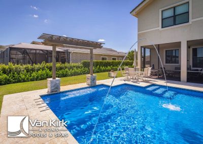 Modern backyard pool with water feature and patio seating, surrounded by greenery.