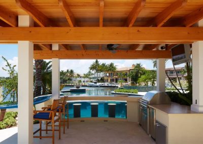 Luxury outdoor kitchen with pool, bar, and waterfront view under a wooden pergola, surrounded by palm trees.
