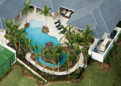 Aerial view of luxury home with tropical pool and lazy river, surrounded by palm trees and lounge chairs, next to tennis court.