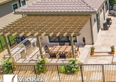 Outdoor pergola with dining area and built-in grill, perfect for backyard entertaining. Van Kirk & Sons Pools & Spas logo.
