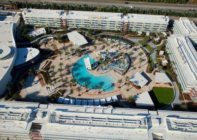 Aerial view of a resort with a large pool, palm trees, and modern buildings surrounded by greenery and a highway.