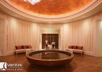 Elegant indoor seating area with round water feature, luxurious lighting, and Van Kirk Commercial Pools & Spas branding.