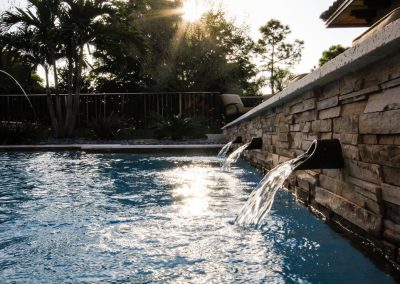 Sunlit pool with stone raised wall and cascading water fountains, surrounded by lush trees. Perfect outdoor relaxation spot.
