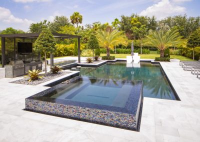 Luxury backyard with a straight pool, hot tub, and pergola in a tropical garden setting under a bright blue sky.