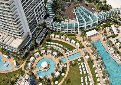 Aerial view of a luxurious resort with pools, gardens, and lounge areas set against modern high-rise buildings.