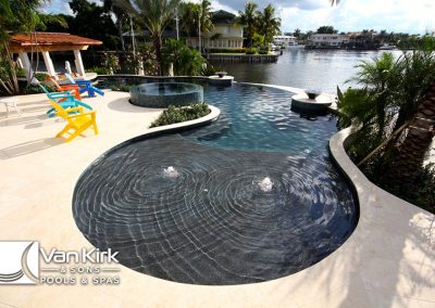 Luxury waterfront pool with spa, colorful chairs, and lush palm trees by Van Kirk & Sons Pools & Spas.