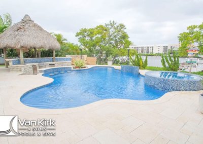 Luxury backyard freeform pool with waterfall and thatched tiki hut by a lake, surrounded by tropical plants.