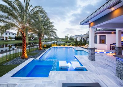 Luxurious outdoor geometric pool area with palm trees, lit by fire features, beside modern homes and a serene lake backdrop.