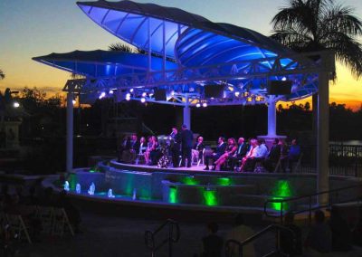 Outdoor evening event at a lit-up modern pavilion with seated audience and colorful lighting against a sunset backdrop.
