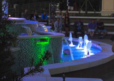 Colorful illuminated fountain with people sitting nearby in the evening.