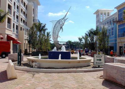 Modern plaza with marlin sculpture fountain, surrounded by buildings and palm trees. Sign reads Keep Right.