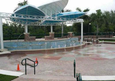 Outdoor water fountain with blue canopy, surrounded by greenery and safety cones, in a recreational area.