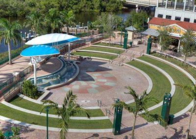 Aerial view of an outdoor amphitheater with circular seating and a nearby river, surrounded by lush greenery and buildings.