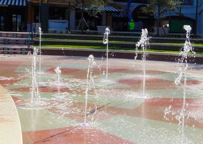 Outdoor plaza with decorative water fountains and colorful tiles, surrounded by trees and modern buildings.