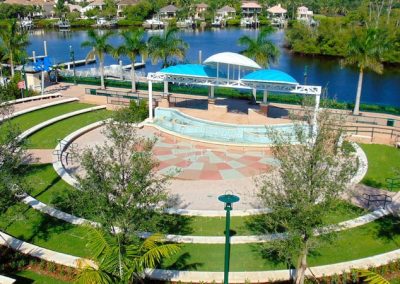 Circular amphitheater with green lawn and covered stage near a river, surrounded by palm trees and residential homes.