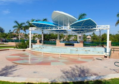 Outdoor stage with blue canopy and water features, set in a sunny park with palm trees.