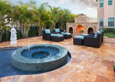 Luxurious outdoor patio with cozy seating, hot tub, and stone fireplace, surrounded by lush greenery and palm trees.