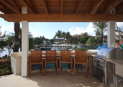 Outdoor kitchen with wooden chairs, a barbecue grill, and waterfront view in a tropical residential setting.