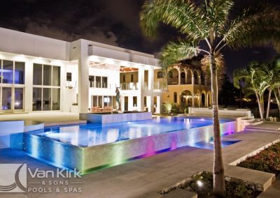 Luxury pool and modern home exterior at night with colorful lighting, palm trees, and a wet edge swimming pool.