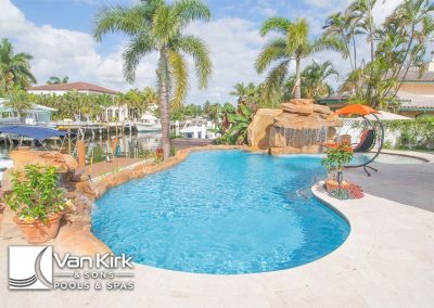 Luxurious backyard freeform pool with waterfall, palm trees, and a view of boats on a sunny day. Perfect for relaxation and leisure.