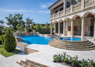 Luxurious villa with elegant columns and a pristine blue lagoon pool surrounded by lush greenery on a sunny day.