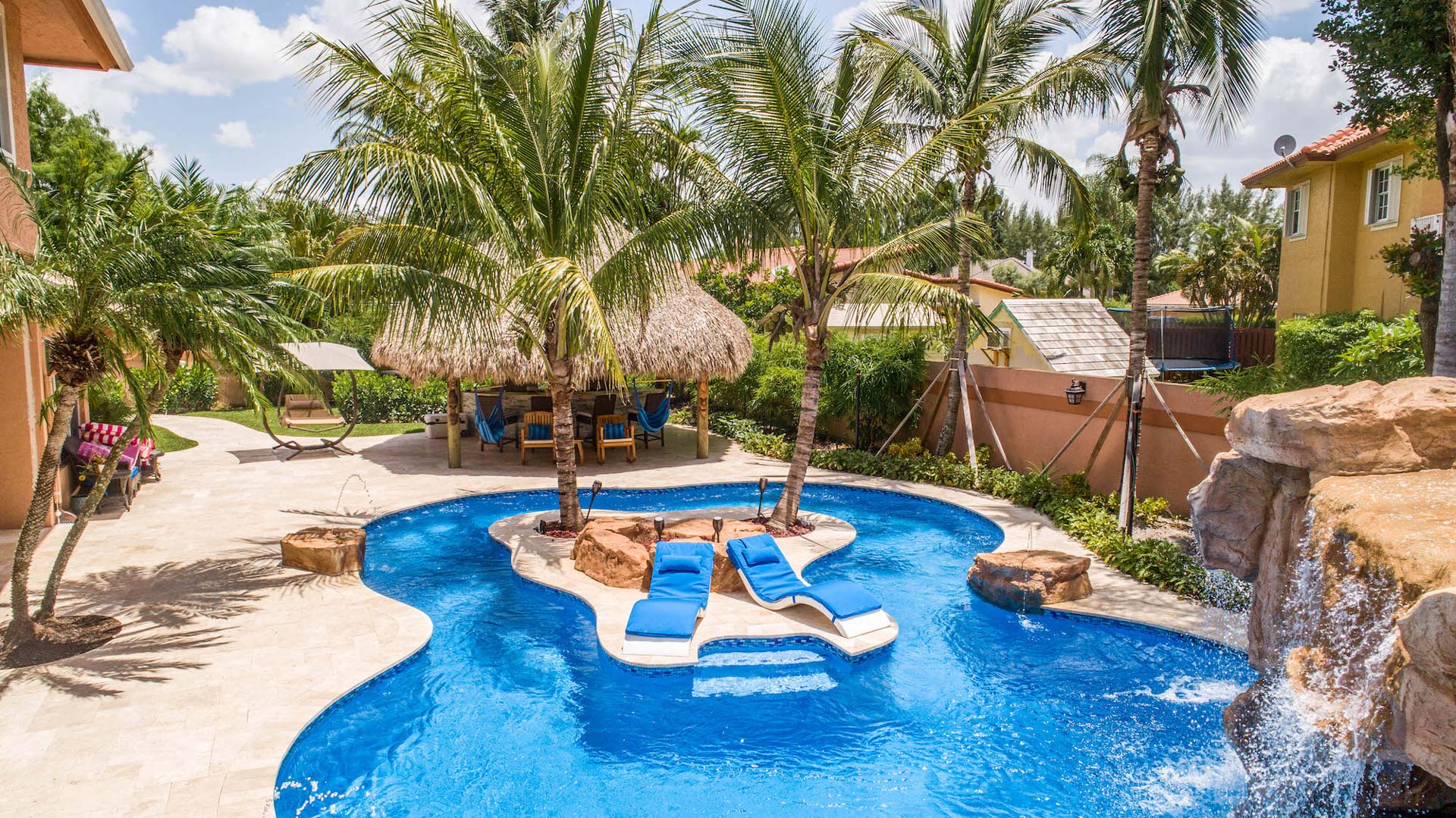 Tropical backyard oasis with a lagoon-style lazy river pool, waterfall, and tiki hut lounge area.