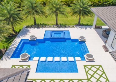 Aerial view of modern luxury pool with geometric design, surrounded by palm trees and a spacious patio.