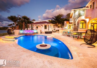Luxurious illuminated freeform style pool and spa with modern design, surrounded by tropical landscaping and a stylish outdoor patio.