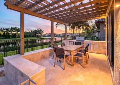 Outdoor kitchen with pergola, dining set, and built-in grill overlooking a serene lake at sunset.
