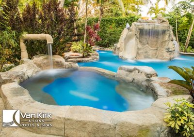 Luxury backyard pool with waterfall feature surrounded by lush greenery and tropical plants.