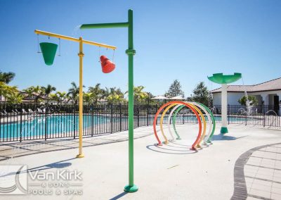 Colorful splash pad with water features near a swimming pool under a clear blue sky.