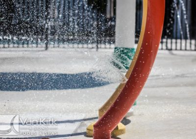 Water splashing from colorful arch at a splash pad, Van Kirk & Sons logo visible.