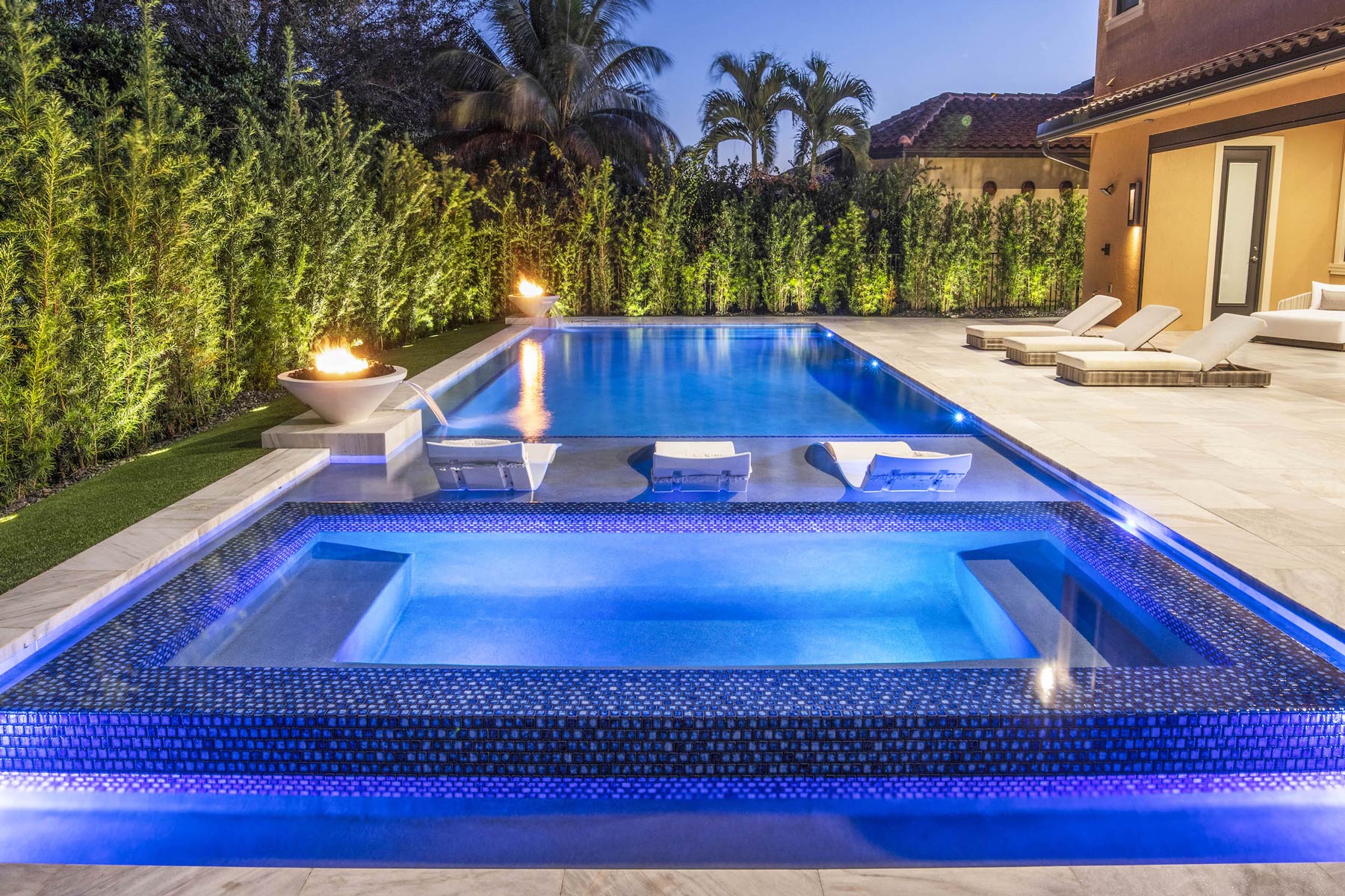 Luxury backyard pool and hot tub with lounge chairs, fire features, and ambient lighting, perfect for evening relaxation.