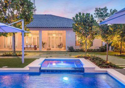 Modern villa exterior with illuminated pool and spa and lush garden at sunset.