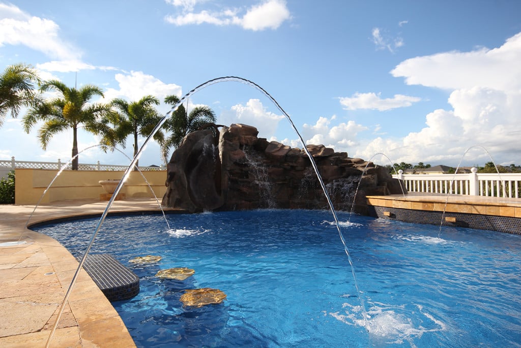 Luxurious backyard pool with deck jets, rock waterfall and fountains under a clear blue sky, surrounded by palm trees.