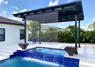 Modern backyard pool with cascading waterfall and pergola under a clear blue sky, bordered by lush greenery.