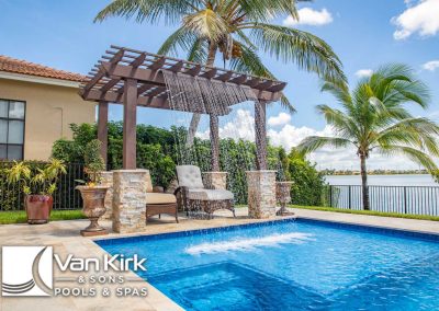Modern backyard oasis with a pool, waterfall pergola, and palm trees overlooking a serene lake.