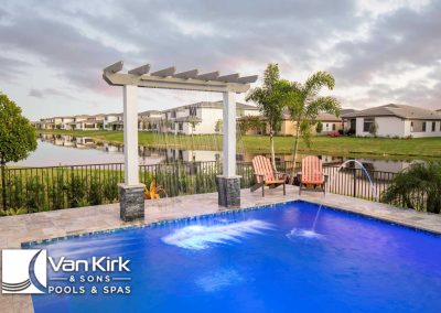 Luxuxrious pool with waterfall from a pergola feature by Van Kirk & Sons Pools & Spas, overlooking serene residential lake view.