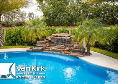 Luxury backyard pool with rock waterfall and lush greenery. Van Kirk logo visible in the corner.