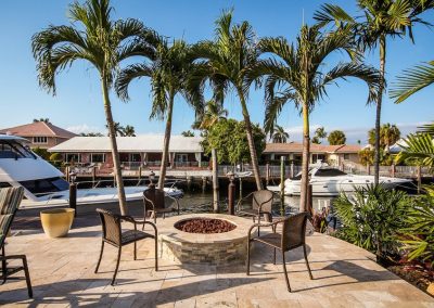 Tropical waterfront patio with fire pit, palm trees, and boats docked nearby on a sunny day.
