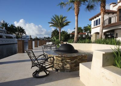 Luxury waterfront patio with palm trees, modern seating, fire pit, and yachts, under a clear blue sky.