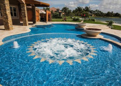 Luxurious backyard pool with fountains and scenic lake view, surrounded by greenery and elegant stone features.