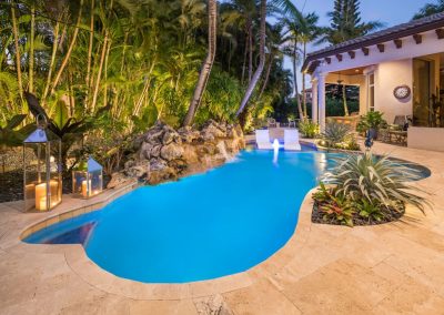 Tropical backyard with illuminated freeform pool, surrounded by lush greenery and cozy patio at dusk. Perfect for relaxation.