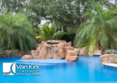 Tropical resort pool with waterfall, surrounded by palm trees and lush greenery, a residential lazy river pool, with Van Kirk & Sons Pools & Spas logo visible.