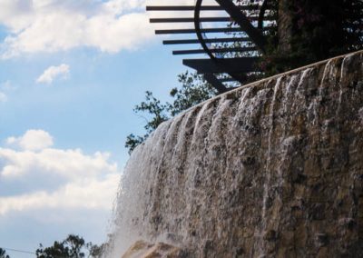 Elegant waterfall with rock design and pergola against a blue sky. Van Kirk & Sons Pools & Spas branding visible.