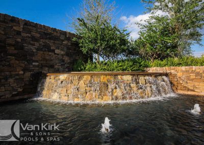 Stone waterfall feature in a pool with cascading water, surrounded by lush greenery and clear blue sky.